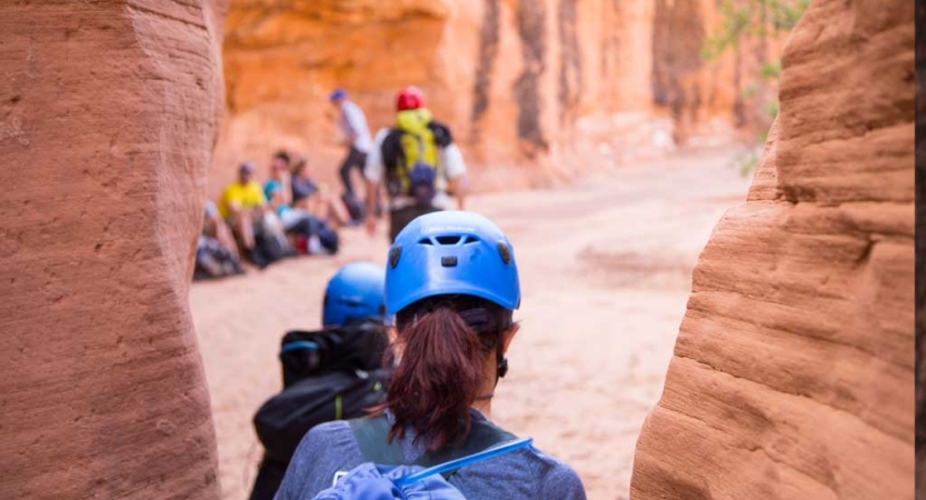From behind, a person wearing a helmet exits a red slot canyon toward a group of others. 